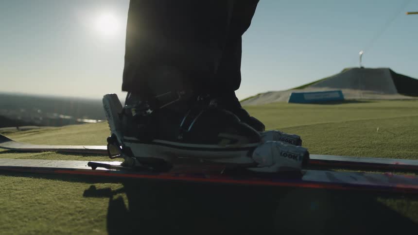 A closeup of ski bindings and boots on grassy terrain with a persons legs visible
