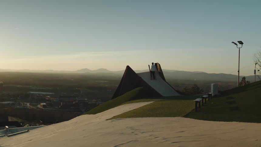 A man carrying skis climbs a ramp built atop a mountain overlooking a valley