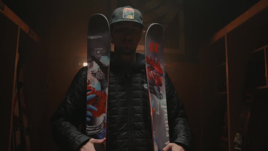 A man holding two snowboards walking forward in a dark environment and he is wearing a cap and warm clothes