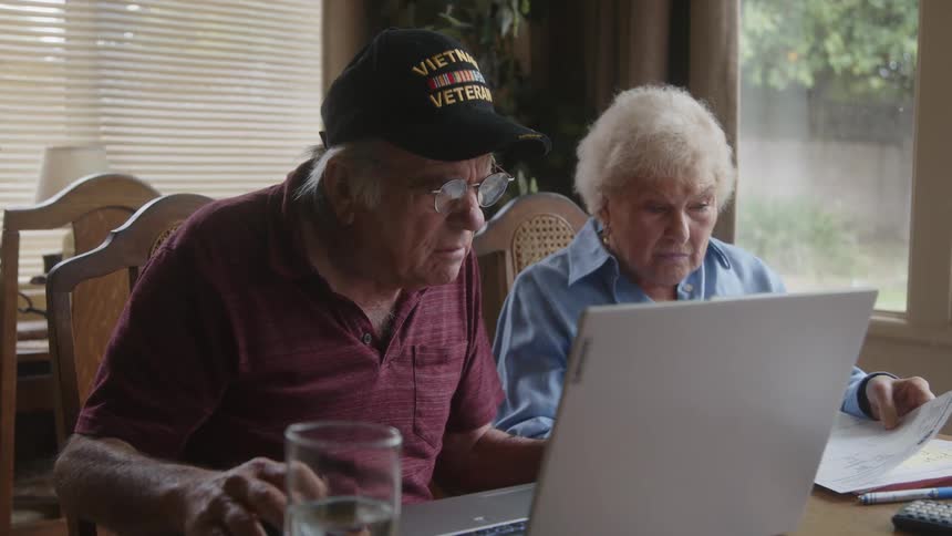 An elderly couple sit at a table with a laptop and papers in front of them