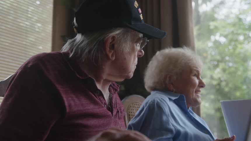An elderly man and an elderly woman are sitting next to each other talking and the woman is showing a paper to the man and they are intrigued
