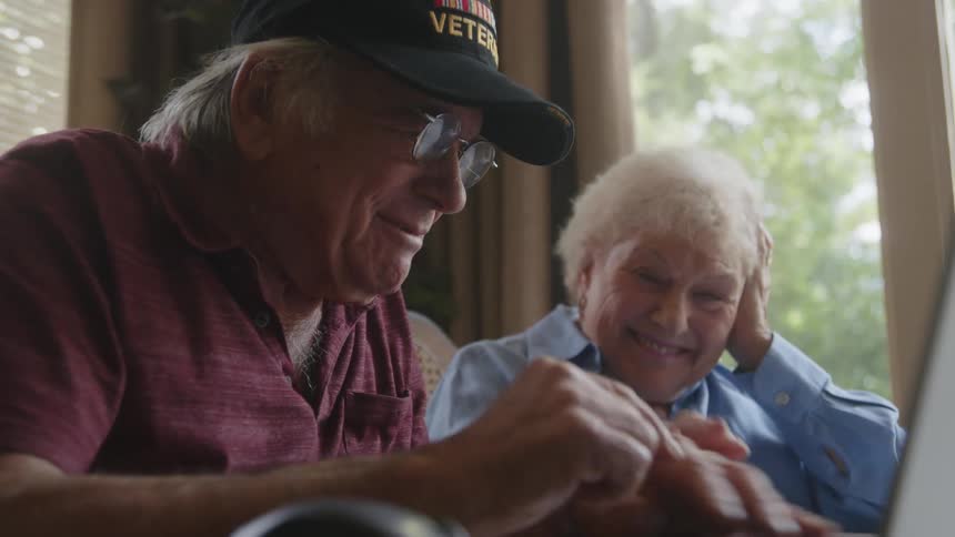 An elderly husband and wife clown around and laugh over a laptop