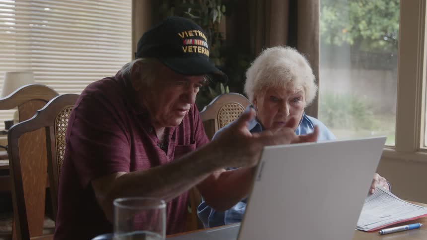 Elderly couple sitting at a table arguing sadly with documents and laptop in front of them