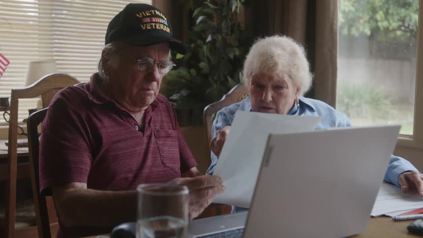 An elderly couple sit at a table with a laptop and papers in front of them