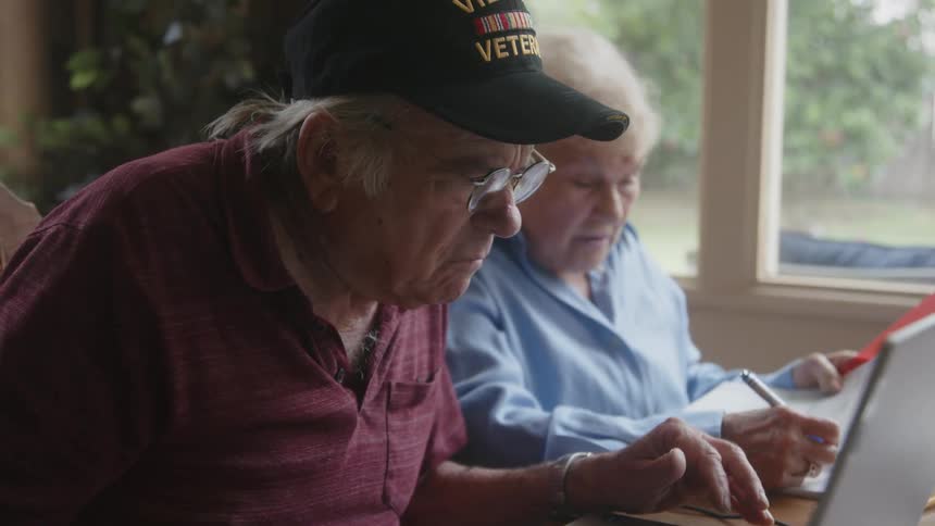 An elderly man in a cap and glasses is working on a laptop in front of him and next to him is an elderly woman who is writing something on a paper