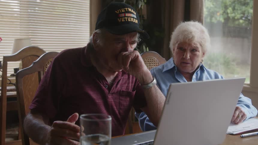 Two elderly people sitting at a table with a laptop talking