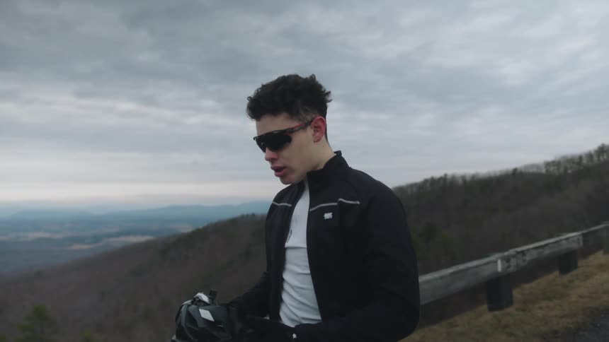 A man takes off a helmet and breathes hard next to a wooden guardrail