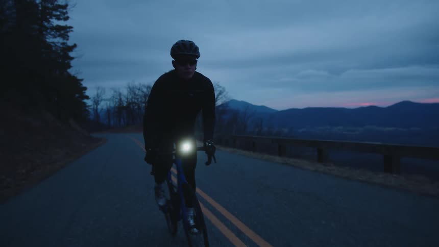 A man rides a bicycle with a headlight on a remote road at dusk