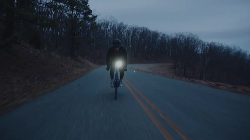 A man rides a bicycle with a headlight down a remote forest road at dusk