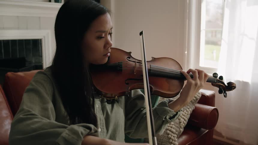 A young asian woman sits on a couch playing a violin