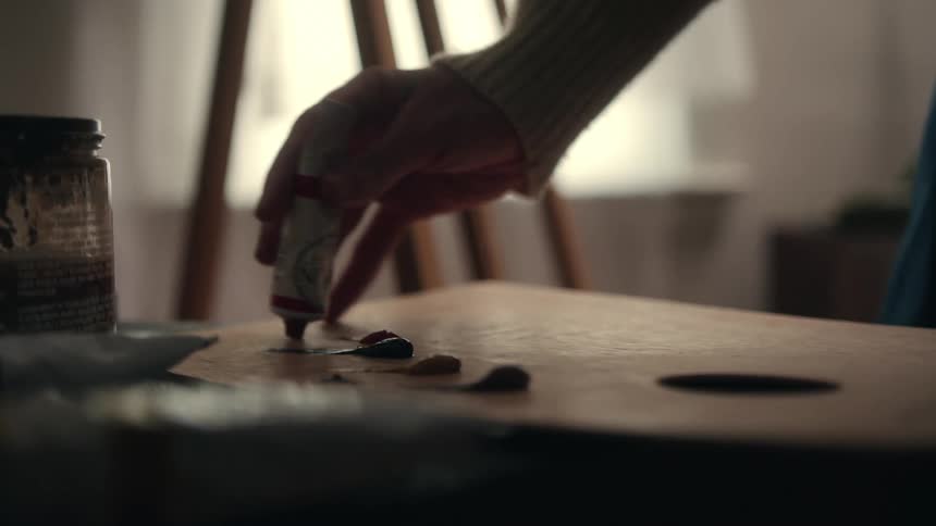 A closeup view of a hand squeezing blue paint from a tube onto a wooden palette which contains several dollops of paint