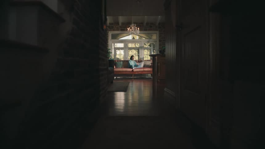 A dimly lit hallway leading to a brightly lit room where a person is seated on a couch