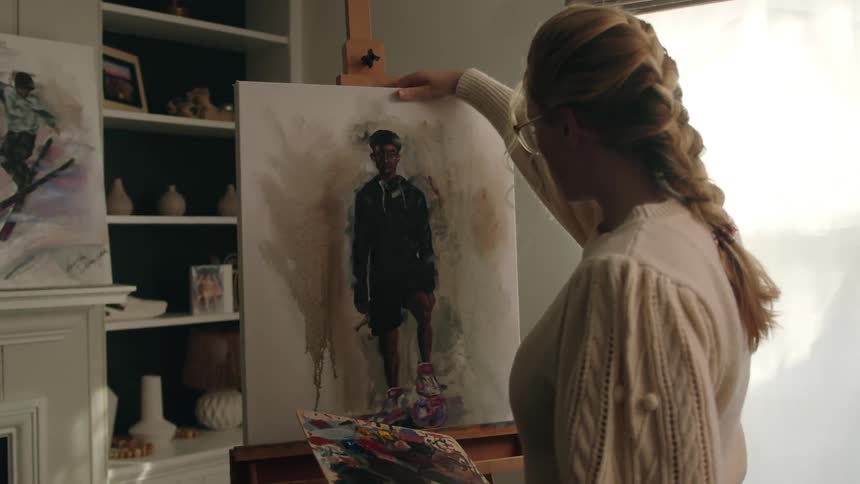 A blonde woman picks up a painting and sets it on an easel