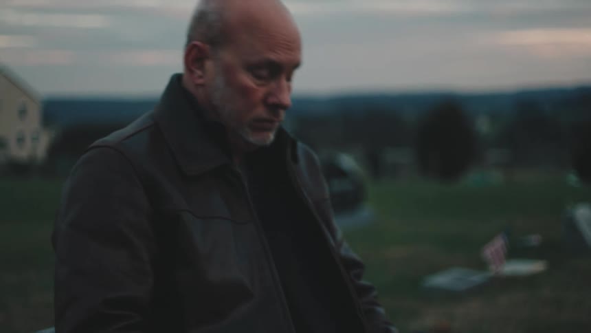 A middleaged man sits in front of a grave in a cemetery during dusk