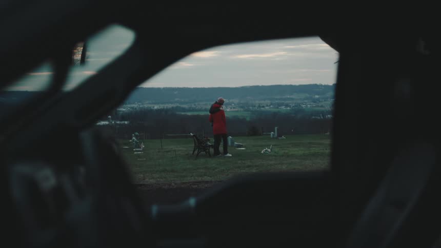 Two individuals stand in an open field with scattered items lie on the ground around them seen from inside a vehicle