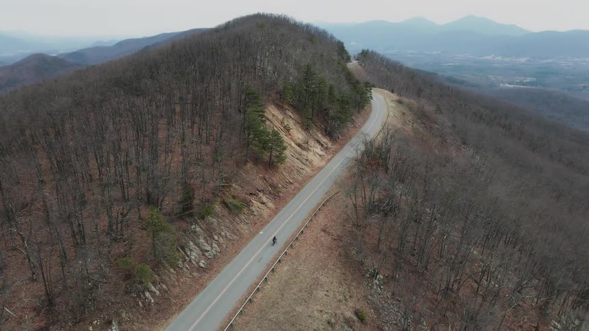 An aerial view of a person riding a bicycle on a winding road cutting through a barren forest