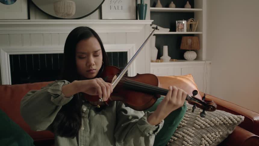 An asian woman draws a bowstring across a violin in a living room