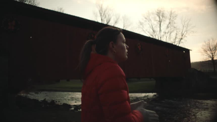 A woman in a red jacket stands next to a bridge over a river against the backdrop of trees and a sunset