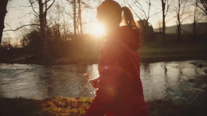 A woman in a red jacket holding a dry white rose walks near a river against the backdrop of trees and a sunset