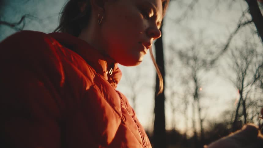 A woman in a red jacket holding a dry white rose against the backdrop of trees and a sunset
