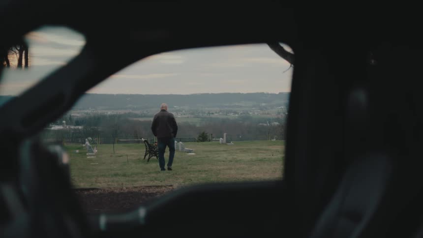 View through a car of a man walking into a cemetery and sitting on a bench