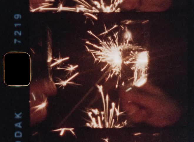 A couple or two young people clink champagne glasses filled with bubbly sparkling wine and the woman drinks from hers while smiling as they hold firework sparklers in their hand celebrating the holidays