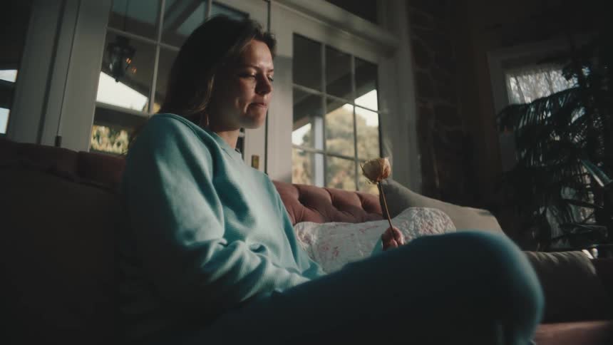 A young woman holding a flower looks sad as she sits on a couch