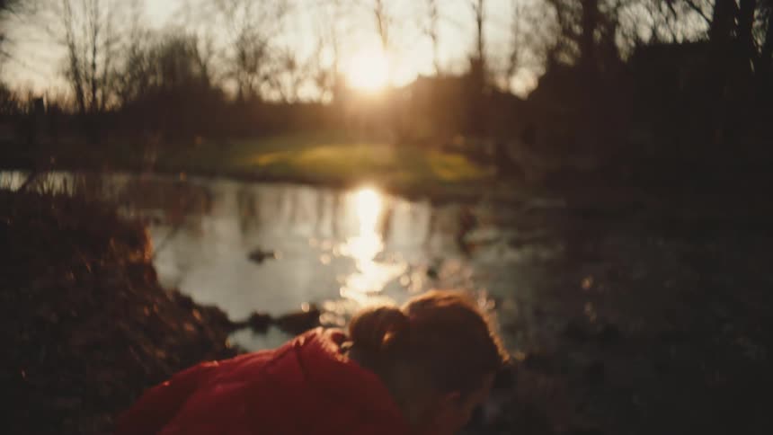 Woman holding a white rose and crouching next to a river with a sun on the horizon