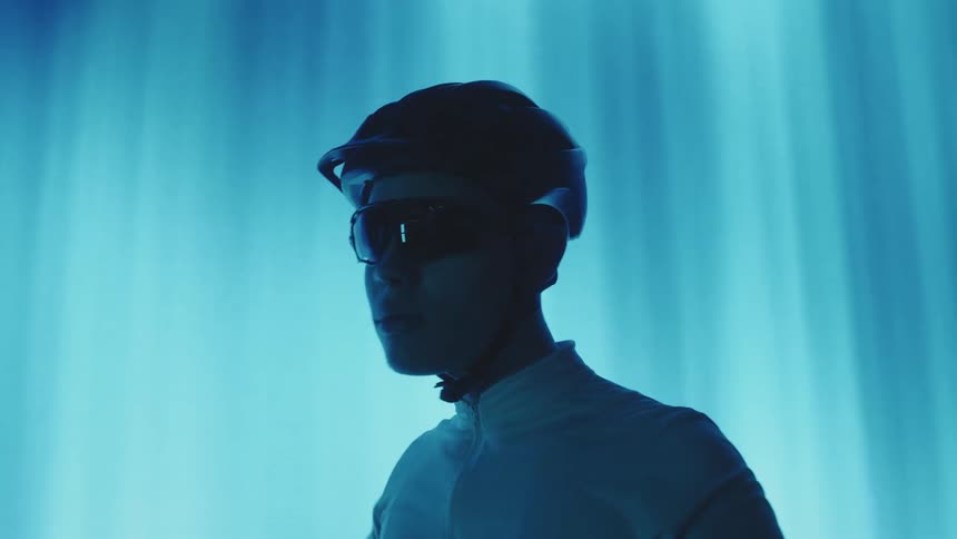 Cyclist man with helmet putting on glasses while indoors with low lighting and blue light in the background