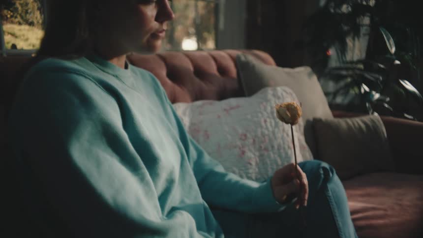 A sad woman holds a flower while sitting on a couch in a living room