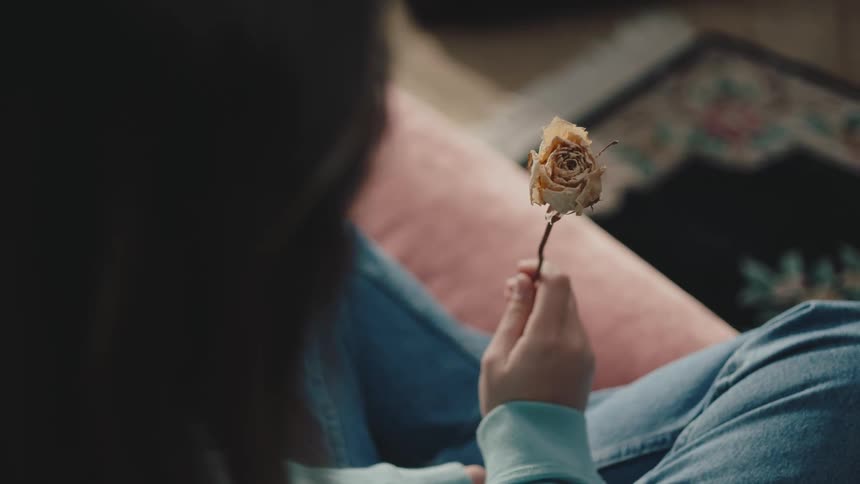 An woman in blue clothes holds a dried rose examining it closely sitting on a couch at home