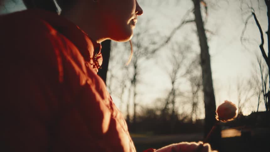 A woman in a red jacket holding a dry white rose against the backdrop of trees and a sunset