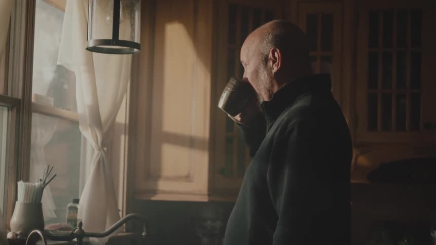 A bald man stands in a kitchen sipping out of a cup