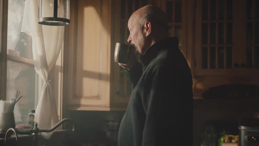 An older man takes a sip from a cup in a kitchen