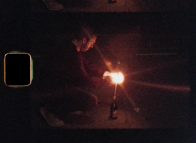 Young man crouching in the street at night lighting a firework rocket that launches into the sky as he stands up and lays his arm around his girlfriend while they watch the sparkles explode on vintage Super 8 film footage