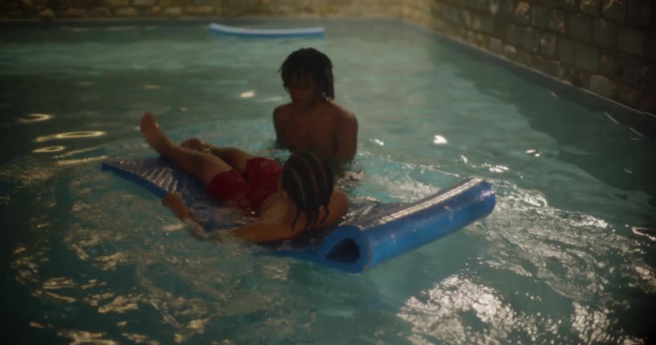 Two boys are in a pool and one of them is lying on a float and the other drops him