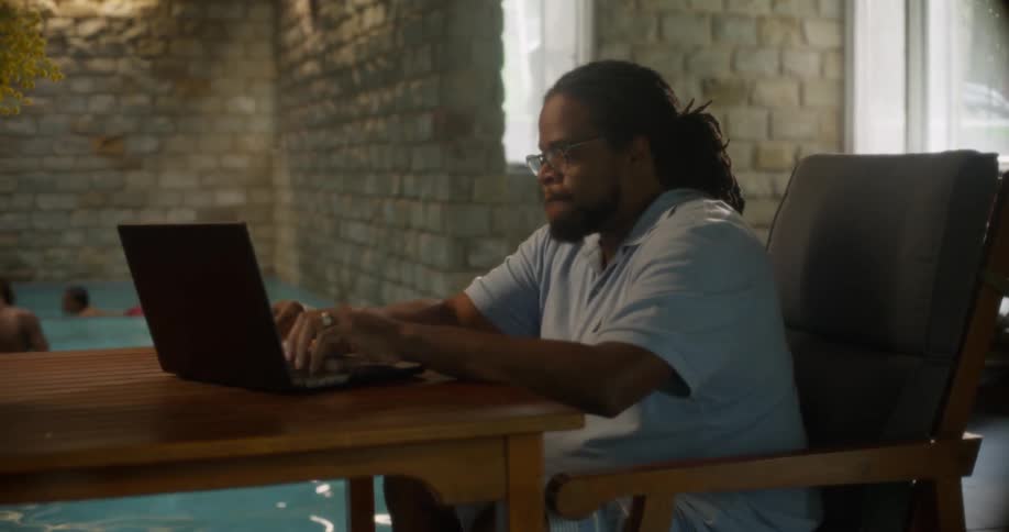 Man working on a laptop and in the background a swimming pool with children playing