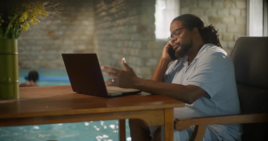 Two kids play in a pool as an adult types on a laptop and answers a smartphone at a table