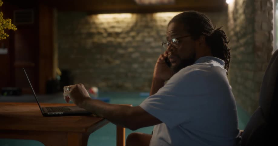 A man is talking on the phone and using his laptop by the pool
