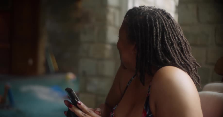 A happy woman holding a smartphone laughing while her children play in the pool