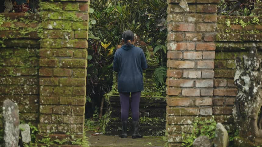 A woman standing in a lush tropical garden alcove surrounded by old brick walls