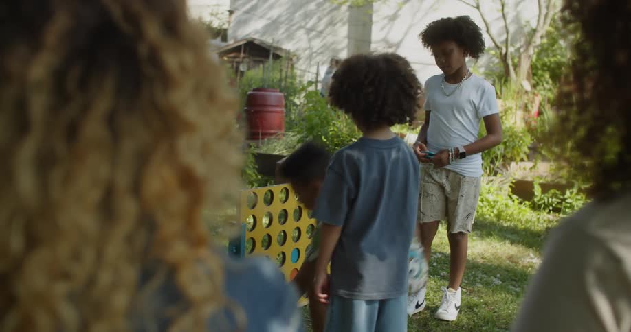 Three little boys are playing Connect Four in an urban garden as their mothers observe