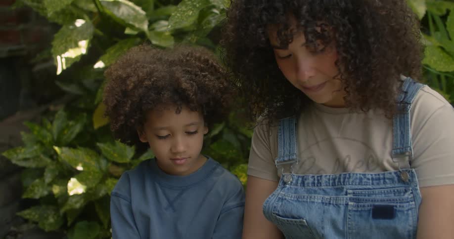 A mother reads to her son from a book while they sit in a garden