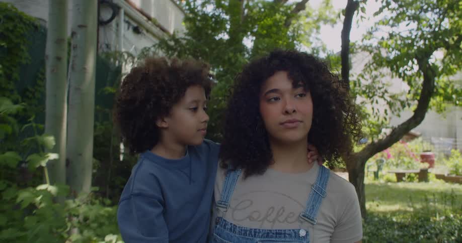 A woman with curly hair is holding her son on her hip while she is walking in a garden