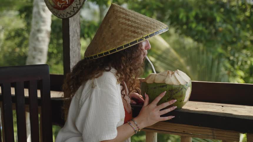 A female tourist on a deck with a cone shaped hat drinks from a coconut