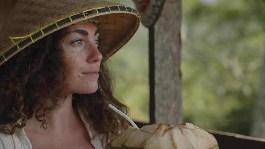 A young woman on vacation in Bali wears a cone shaped hat and sips from a fresh coconut