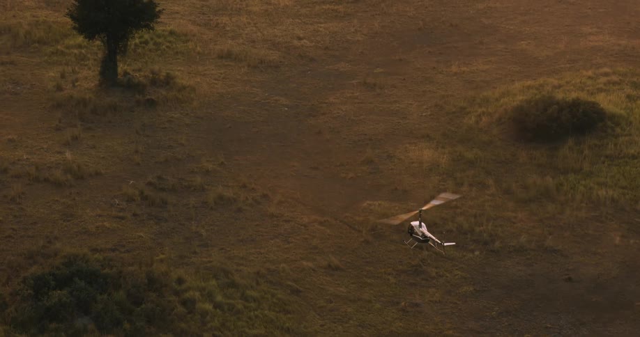 A helicopter with its propellers reflecting sunlight and is flying over an environment with grass on the ground