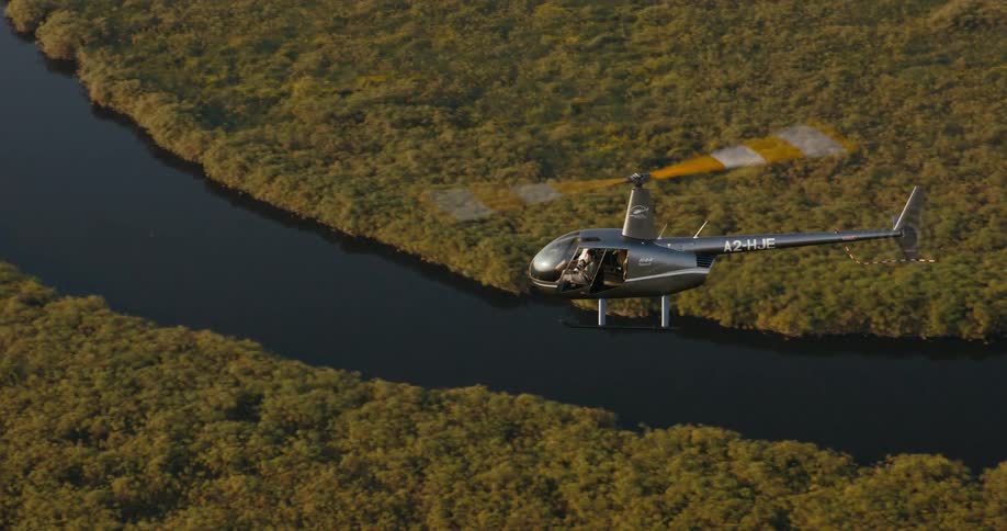 A helicopter flies over a river flanked by dense vegetation on the Okavango Delta