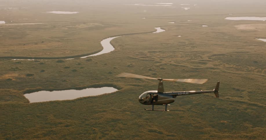 A helicopter flies over a vast grassy plain interspersed with bodies of water