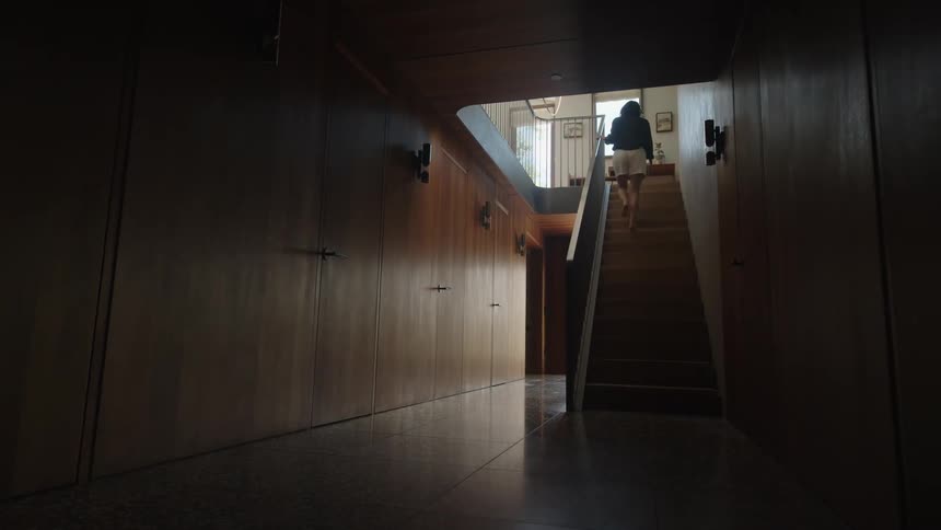 A dimly lit hallway with wooden walls and floors leads to a staircase where a woman is seen walking up